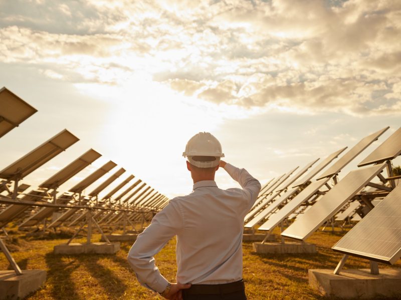 Unrecognizable engineer admiring modern solar farm at sunset Back view of anonymous man in hardhat touching head and looking at modern photovoltaic panels and cloudy sundown sky during work on solar power station